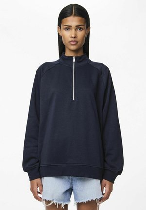 Navy blue sweatshirt with a high collar and half-zip, featuring raglan sleeves and ribbed cuffs, paired with distressed denim shorts.