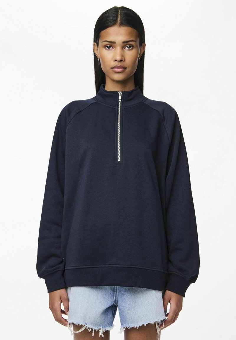 Felpa blu navy con colletto alto e zip a metà, caratterizzata da maniche raglan e polsini a costine, abbinata a pantaloni shorts in denim strappato.