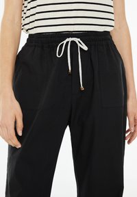 Pantalones negros de cintura elástica con cordón, que presentan detalles en color beige, bolsillos laterales y un tejido ligero y texturizado.