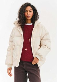 Krémově zbarvená prošívaná bunda s prošívaným vzorem, vybavená vysokým límcem, patentními knoflíky a prostorným střihem, vrstvená přes vínový svetr.