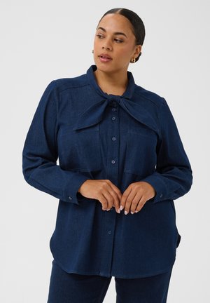 Femme portant une chemise en jean bleu foncé à boutons avec un grand nœud noué au col, debout, les mains jointes à la taille.
