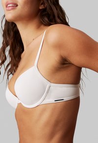 Reggiseno beige chiaro con tessuto liscio, supporto con ferretto e spalline sottili regolabili. Dettagli testurizzati evidenti e piccolo logo sulla fascia.