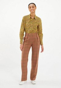 Chemise à manches longues verte à motifs floraux associée à un pantalon taille haute en tartan marron et rouge et des baskets blanches. Tissu lisse et léger.