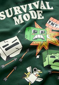 Grüner Stoff zeigt ein "Survival Mode"-Design mit Comicfiguren, Pfeilen und Explosionen, hervorgehoben durch lebendige Farben und kräftige Grafiken.