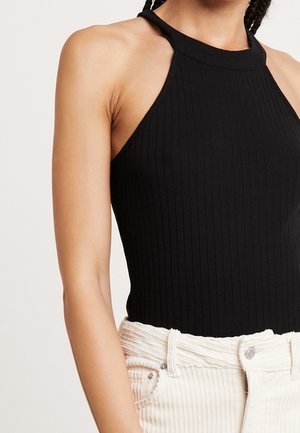Top halter de canalé negro con un escote alto y diseño sin mangas, combinado con pantalones de pana en color blanco roto con detalles de botones y bolsillos.