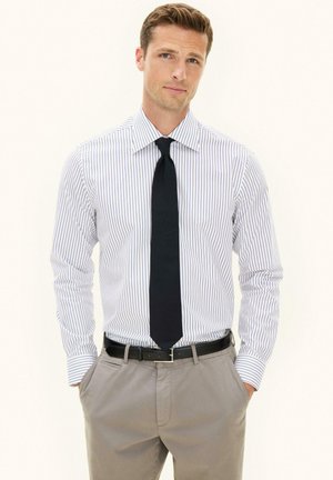 Mężczyzna ubrany w białą koszulę z cienkimi pionowymi paskami, czarny krawat, beżowe spodnie i czarny pasek, stojący z rękami w kieszeniach.