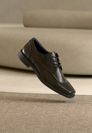 Pantof elegant de piele neagră, cu șireturi, plutind deasupra unui covor bej texturat, cu un fundal de canapea bej asortată.