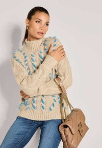 Frau in einem beigen grob gestrickten Pullover mit blauen Details, blauen Jeans, goldenen Kreolen und einer hellbraunen Ledertasche mit goldener Kettenriemen.