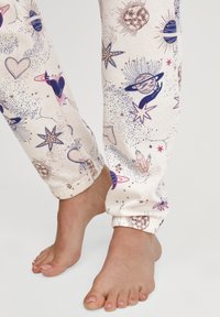 Lichte pyjamabroek in crème met een kosmische print van harten, planeten, sterren en kristallen. Met een manchet bij de enkels.