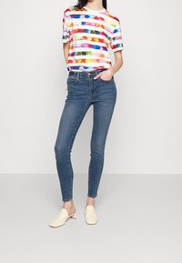 Camiseta de rayas colorida con mangas cortas, combinada con jeans ajustados azul y mules de tacón bloque beige, creando un atuendo informal.