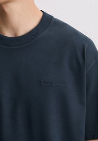 Marineblaue Rundhals-T-Shirts mit dezentem "Marc O'Polo"-Logo auf der linken Brust, getragen von einer Person vor einer hellen strukturierten Wand.