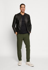 Schwarze Leder-Bomberjacke, schwarzes T-Shirt, grüne Hose und weiße Sneakers. Die Jacke verfügt über einen Reißverschluss und gerippte Details.