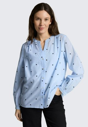 Młoda kobieta o długich brązowych włosach, ubrana w jasno niebieską bluzkę na guziki z małymi czarnymi wzorami serc oraz czarne spodnie, z jedną ręką w kieszeni.