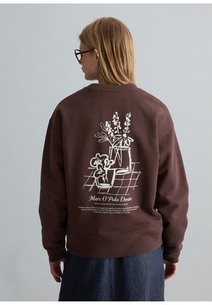 Persona con gafas y cabello rubio lleva una sudadera marrón con un dibujo lineal floral blanco y el texto "Marc O'Polo Denim" en la espalda.
