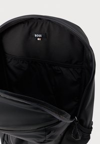 Melna mugursoma ar atvērtu galveno nodalījumu, kurā redzama iekšējā kabata un pie rāvējslēdzēja atvēruma iesieta "BOSS" birka.