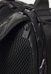 Rucsac negru cu un panou din plasă texturată, material impermeabil elegant și un fermoar distinctiv cu detalii ale logo-ului.