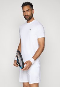 Λευκό κοντομάνικο μπλουζάκι με μικρό πράσινο λογότυπο, συνδυασμένο με λευκές βερμούδες. Κρατάει μια μαύρη ρακέτα τένις, φορώντας λευκά περιβραχιόνια.