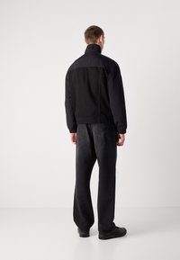 Una persona in piedi di spalle che indossa una giacca nera, jeans neri larghi e sneaker nere su uno sfondo bianco uniforme.