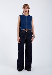 Top sin mangas de denim recortado en azul oscuro con dobladillo sin terminar, combinado con unos pantalones vaqueros de pierna ancha de tiro alto en azul oscuro. Zapatos negros y pulsera de plata.