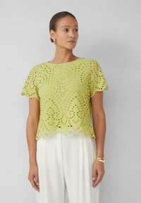 Zielona koronkowa bluzka z krótkimi rękawami, falbaniastym dołem i misternymi wzorami wycięć, w zestawie z białymi spodniami i złotą bransoletką.