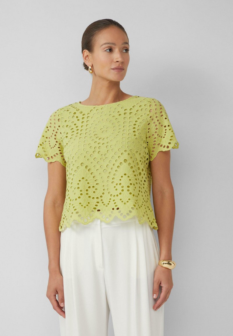 Zielona koronkowa bluzka z krótkimi rękawami, falbaniastym dołem i misternymi wzorami wycięć, w zestawie z białymi spodniami i złotą bransoletką.