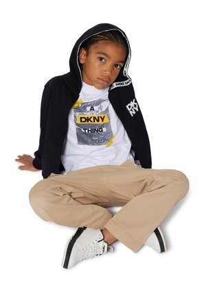 Black zip hoodie with white accents, white graphic t-shirt featuring yellow text, beige pants, and white sneakers with black detailing.