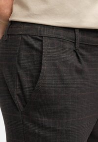 Mørke ternede bukser med en let glans, en struktureret pasform, forreste læg og subtile burgunderrøde accenter mod en sort baggrund.