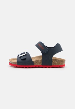 Navyblaue Sandalen mit rotem Gummisohle, Korkfußbett und verstellbaren Riemen mit einer silbernen Schnalle. Strukturierte Oberseite und gezackter Sohlenrand.