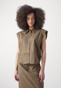 Camicia marrone a maniche corte con colletto, realizzata in tessuto texturizzato e con bottoni frontali. Indossata con una gonna abbinata, creando un outfit coordinato.