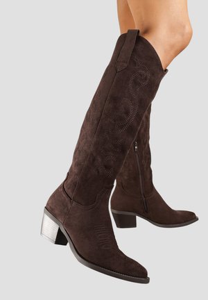 Bottines en daim marron foncé, hauteur genou, avec des coutures décoratives, une fermeture éclair sur le côté et des talons en bloc argentés épais, portées par une personne sur un fond gris.