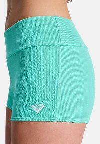 Shorts vert menthe avec une texture côtelée, une taille haute et un petit logo en forme de cœur blanc près de l'ourlet. Couleur unie sans motifs.