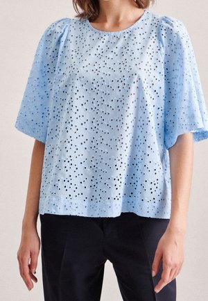 Femme portant un chemisier bleu clair à manches courtes avec un motif ajouré, associée à un pantalon noir, debout devant un fond uni.