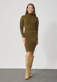 Oliwkowo zielona, prążkowana dzianinowa sukienka z wysokim kołnierzem i dopasowaną sylwetką, w parze z beżowymi kozakami za kolano. Gładka tekstura i jednolity kolor.