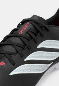 Μαύρο ποδοσφαιρικό παπούτσι κατασκευασμένο από υφή συνθετικό υλικό, με κόκκινες λεπτομέρειες και λευκό γραφικό σχέδιο στην πλευρά.