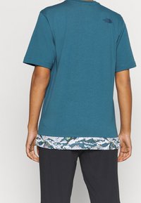 Turkusowy t-shirt z krótkim rękawem z kontrastowym, górskim wzorem na dolnym zakończeniu. Materiał wydaje się być miękki; posiada logo na górnej części pleców.