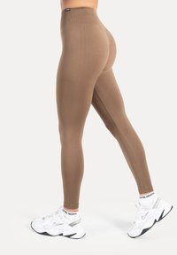 Leggings marron taille haute, à texture lisse, coupe ajustée et design sans coutures, associés à des chaussures de sport blanches et des chaussettes à motifs.