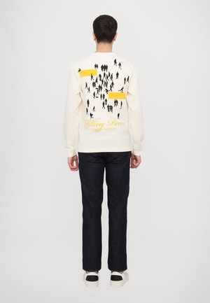 Long-sleeve cream shirt featuring a black graphic of people and a yellow accent on the back. Paired with dark denim pants and white shoes.