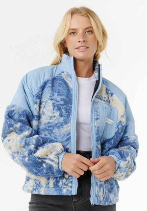 Frau in einer hellblauen und weißen gemusterten Fleecejacke über einem weißen Oberteil, die den Reißverschluss der Jacke mit beiden Händen anpasst.