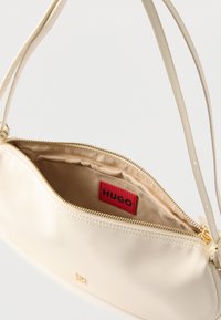 Τσάντα ώμου σε κρεμ χρώμα με χρυσό φερμουάρ, ανοιχτή για να φαίνεται το μπεζ εσωτερικό και η κόκκινη ετικέτα με το κείμενο "HUGO" στο εσωτερικό.