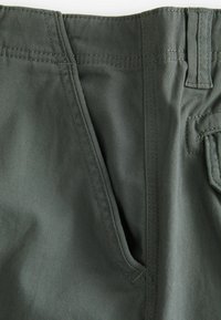 Dunkelgrüne Stoffhosen mit glatter Textur, scharfen Falten und verstärkten Nähten entlang des Bundes und der Taschen.