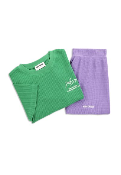 Camicia verde a maglia waffle con grafica bianca di coccodrillo e pantaloni viola a maglia waffle, entrambi etichettati "Bobo Choses", piegati su sfondo bianco.