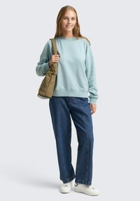 Világoskék pulóver bordás textúrával, laza szabással és ejtett vállakkal, kék bőváló farmerral és fehér tornacipővel párosítva.