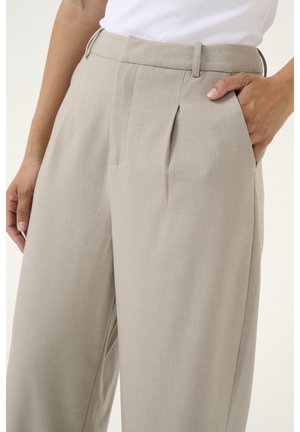 Personne portant un pantalon plissé beige avec des poches latérales, une main reposant à l'intérieur de la poche et un haut blanc rentré.