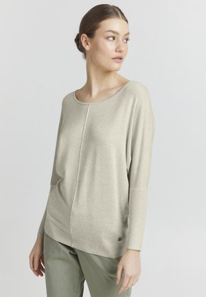 Lys beige strikkegenser, vid scoop-hals, lange ermer, ribbede mansjetter, senter søm-detalj, parret med olivengrønne bukser.