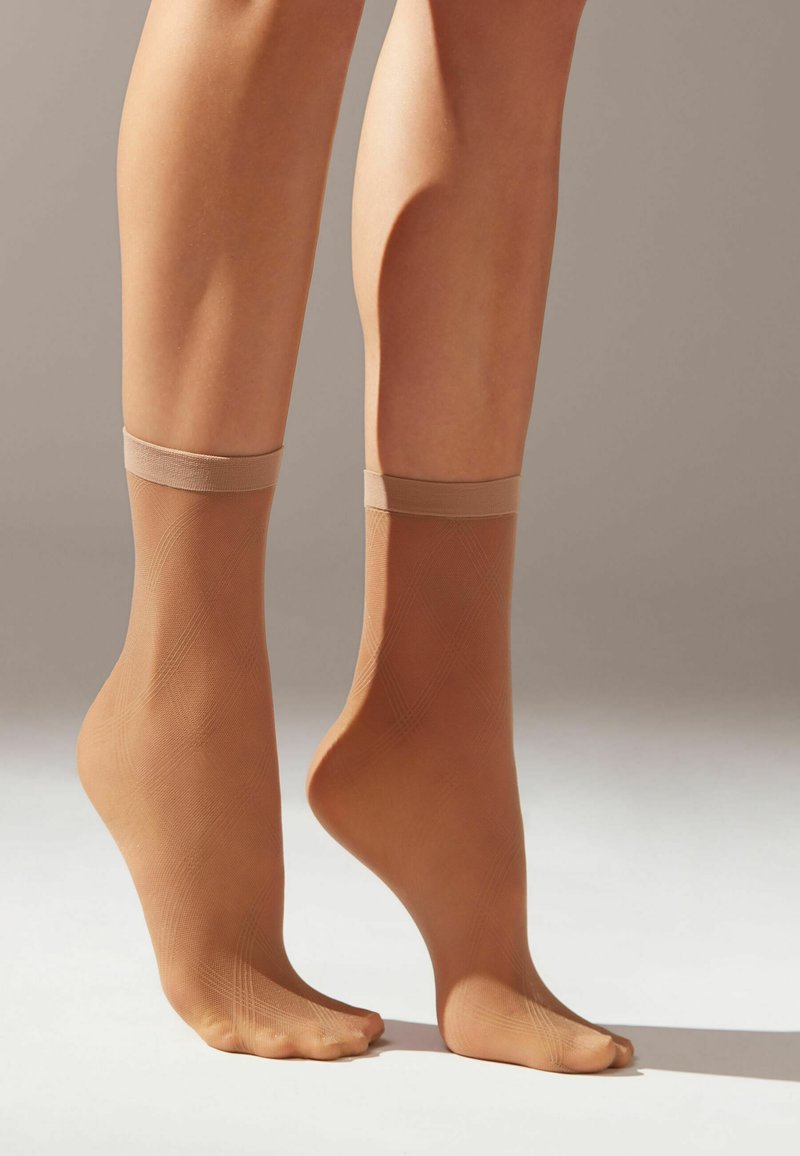 Beige ankelstrumpor med en slät, genomskinlig textur och ett lätt, elastiskt band upptill. Mönstrade med subtila diagonala linjer.