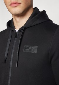 Felpa nera con zip realizzata in tessuto morbido, caratterizzata da un patch logo testurizzato e dettagli con stringhe. Design aderente con tasche a marsupio.