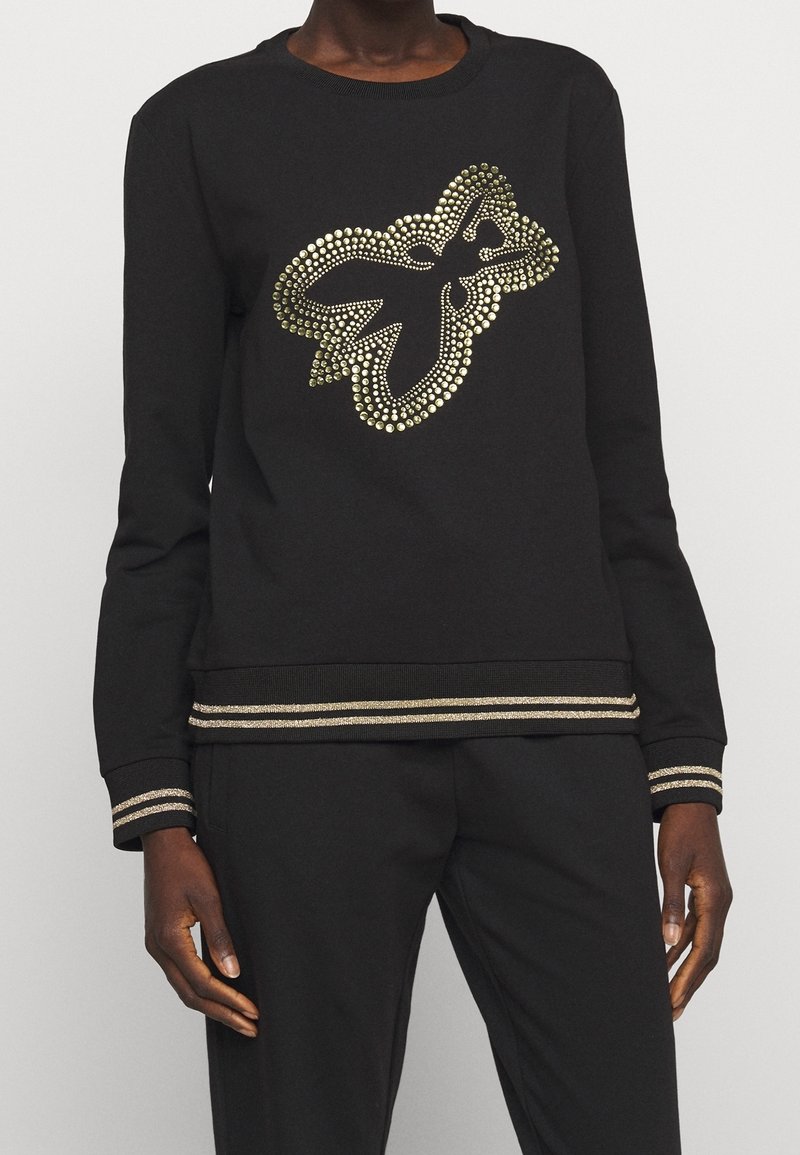 Haut noir à manches longues avec un motif de papillon orné de clous dorés sur la poitrine et des bandes dorées assorties aux poignets et à l'ourlet, porté avec un pantalon noir.