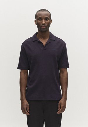 Mężczyzna ubrany w ciemnogranatową koszulkę polo z krótkim rękawem, stojący prosto na jednolitym jasnoszarym tle.