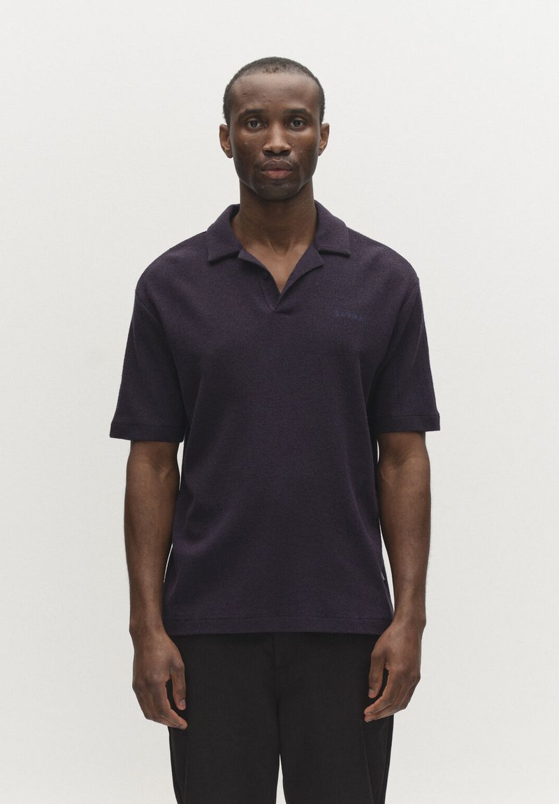 Uomo che indossa una polo a maniche corte blu navy scuro, in piedi dritto contro uno sfondo uniforme grigio chiaro.