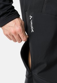 Pantalones deportivos negros con un bolsillo lateral con cremallera. Hechos de un material elástico, que presenta un ajuste ceñido y una mínima marca que muestra "Vaude."